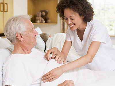 Nurse Helping Senior Man Into Bed In Hospital Ward